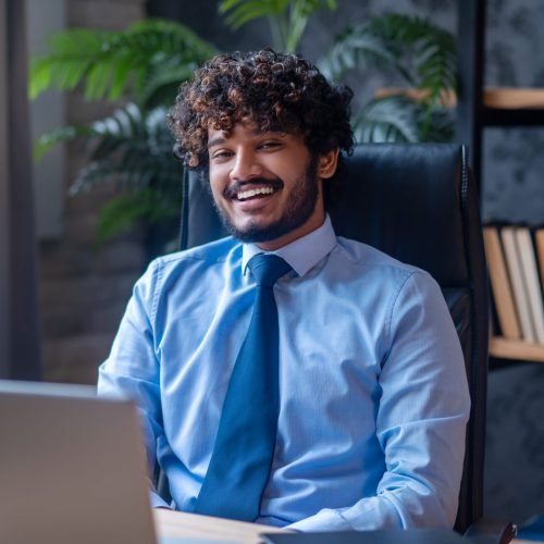 Good mood. Indian attractive young curly haired man with toothy smile in blue shirt and tie sitting at office desk looking at camera